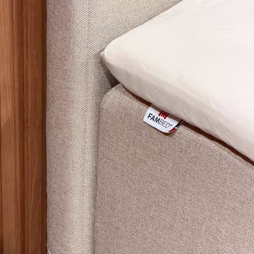 Lähikuva sängyn kulmaa peittävästä beigestä päällisestä, jossa on FAMBED-merkki ja jonka päällä on valkoinen lakana